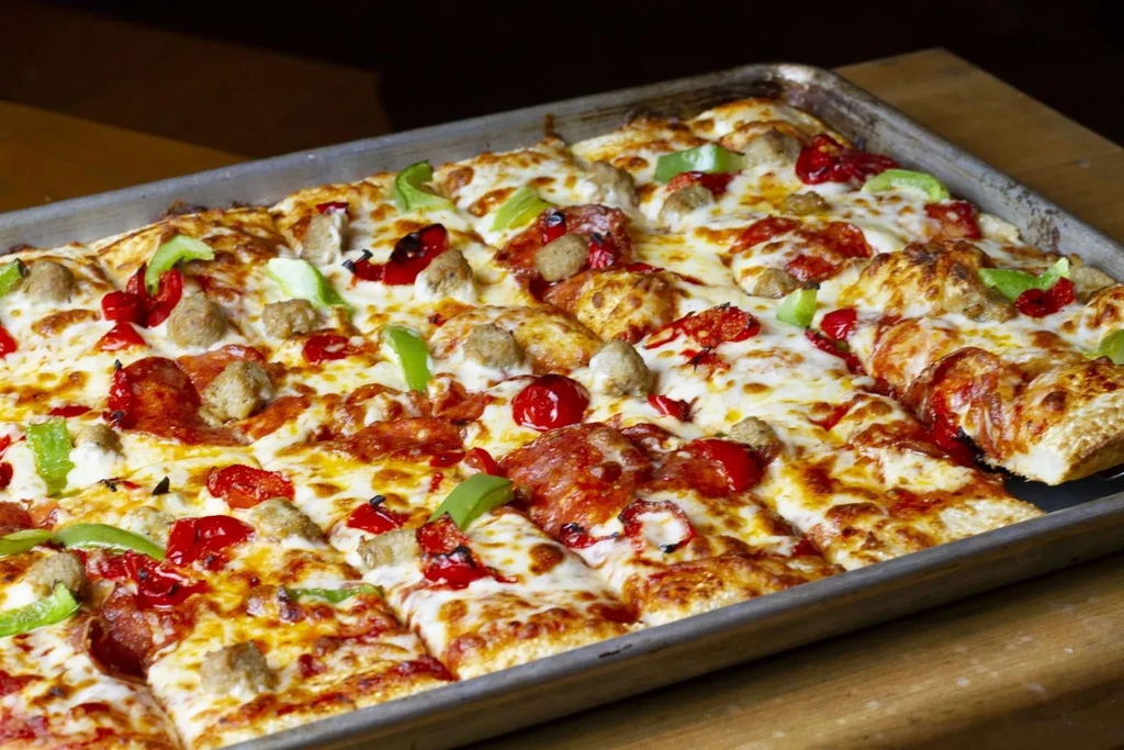 Rectangular deep-dish pizza in tray with cheese, meats, and vegetables.