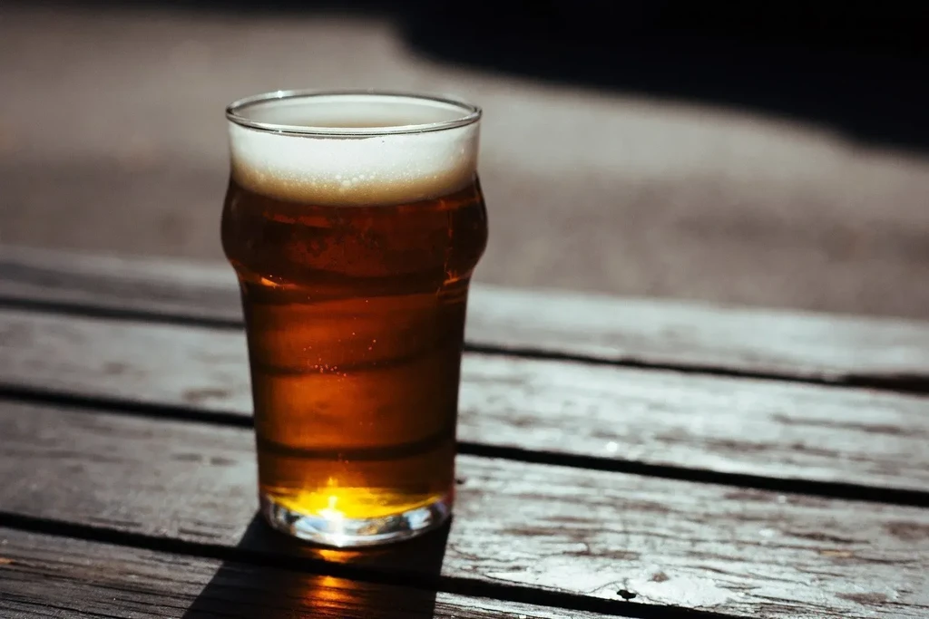 Pint glass of amber beer on a sunny rustic wooden table.