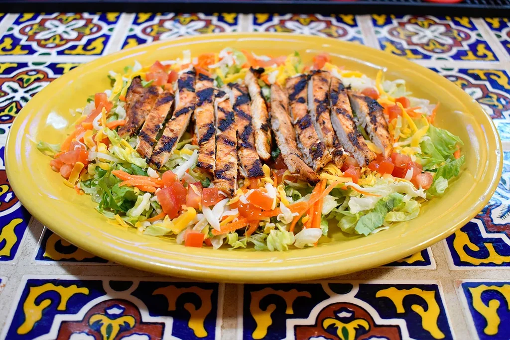 Grilled chicken salad on a yellow plate with colorful tiled background.
