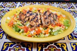 Grilled chicken salad on a yellow plate atop colorful mosaic tiles.