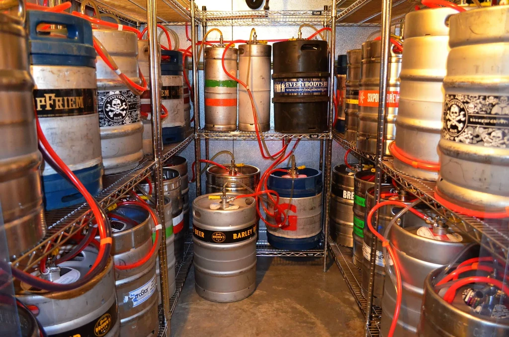 Crowded beer cellar with various branded kegs on shelves connected by tubing.