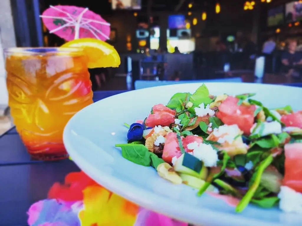 Colorful salad, tiki drink, and lei with blurred restaurant background.