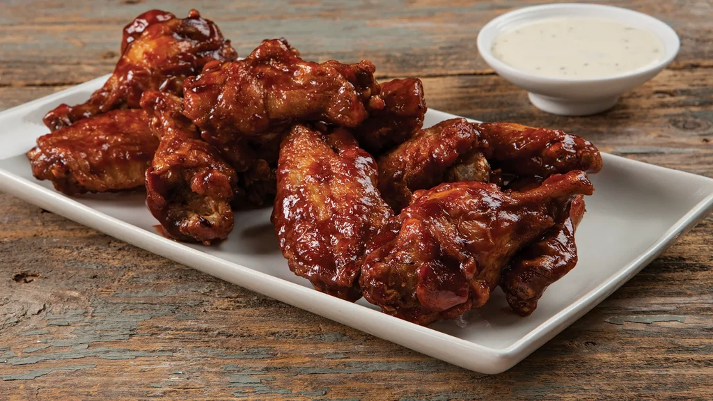 Reddish-brown glazed chicken wings, dipping sauce, on a rustic wooden surface.