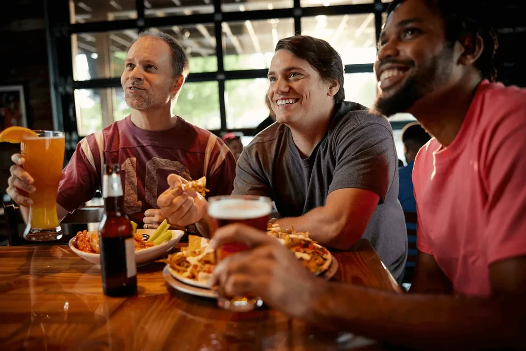 Three men enthusiastically watching, eating, and drinking at a bar.