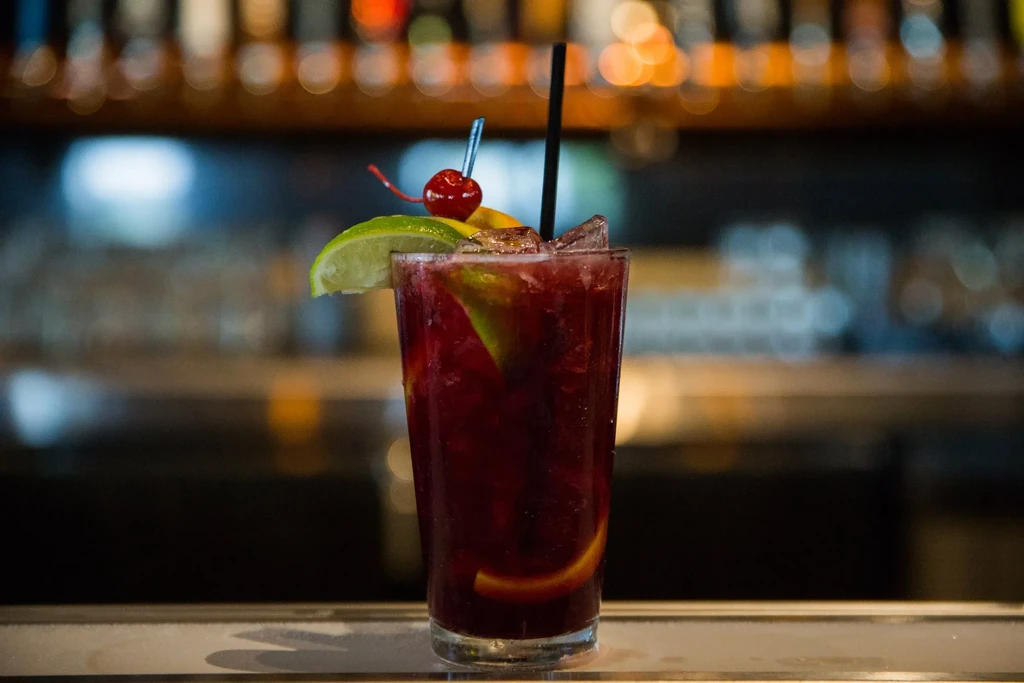 Tall glass of red cocktail with lime, orange, and cherry.