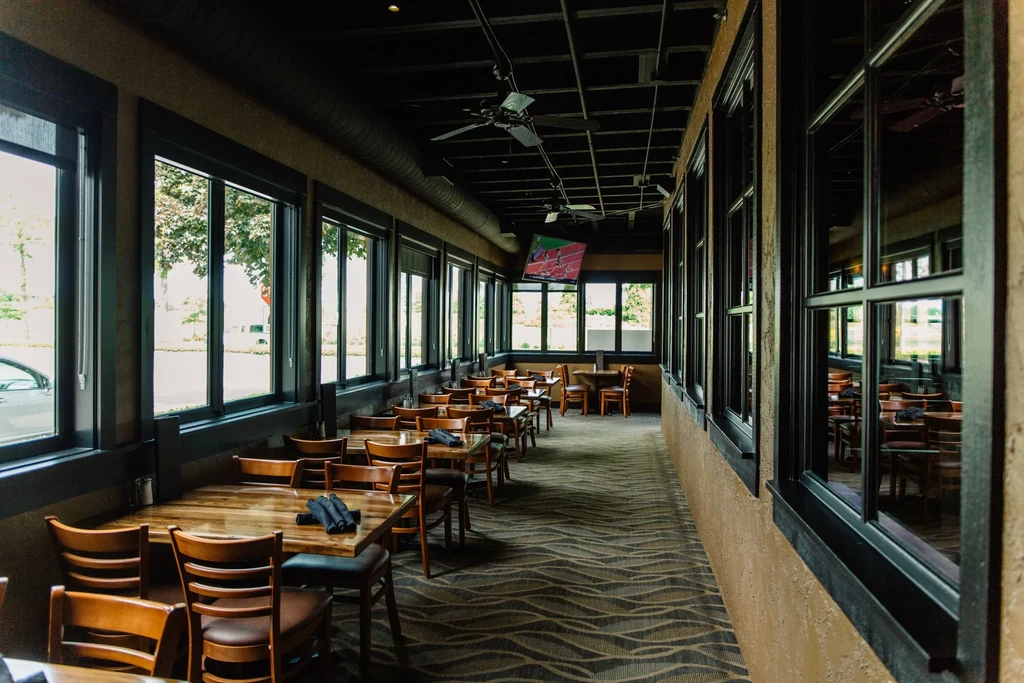 Restaurant sunroom with wooden tables, large windows, and dark ceiling.