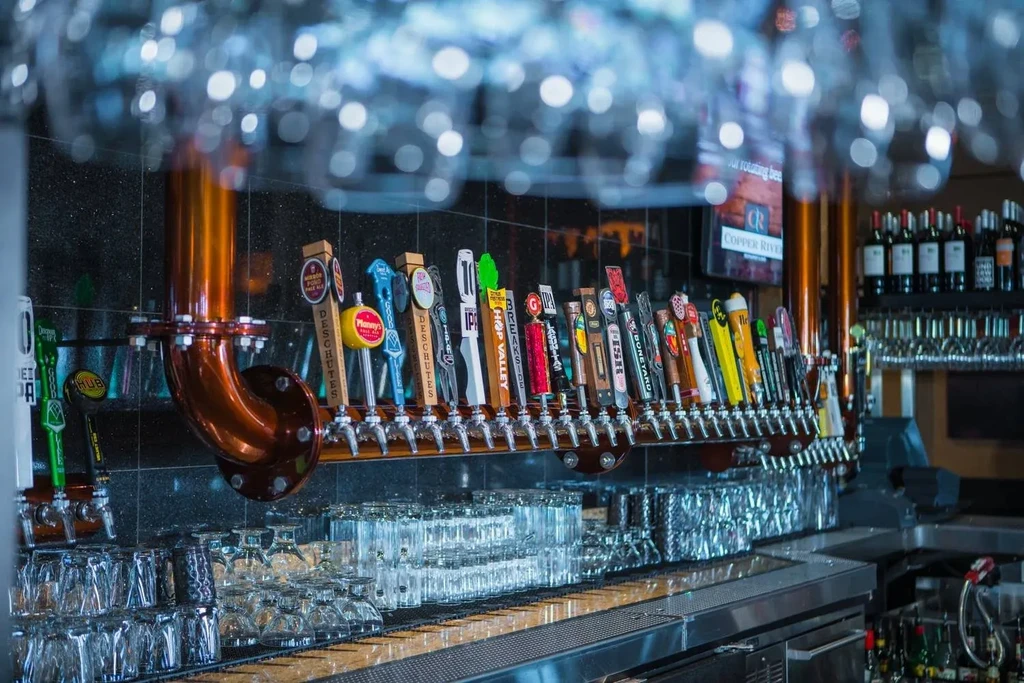 Row of colorful beer taps, prominent copper piping, and glassware at a bar.