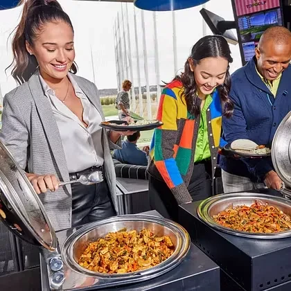 Three smiling people serving buffet food at a TopGolf driving range.