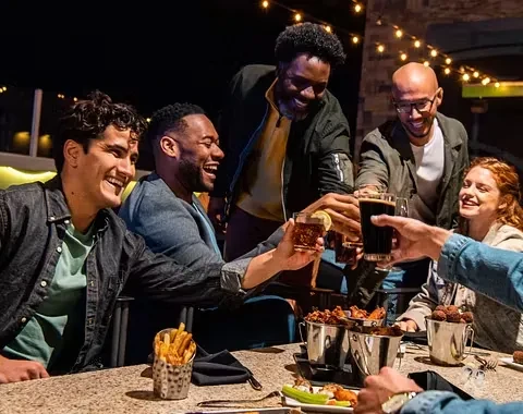 Five diverse friends smiling, toasting drinks and eating food outdoors.