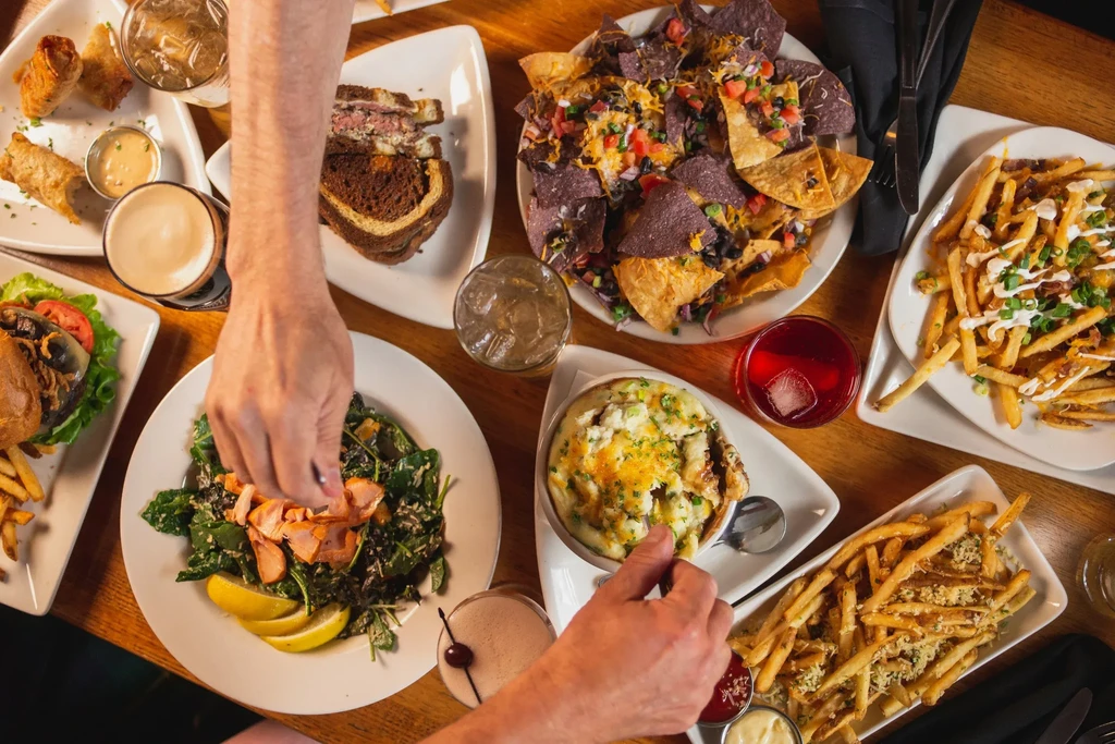 Overhead: wooden table with diverse food, drinks, and hands dining.