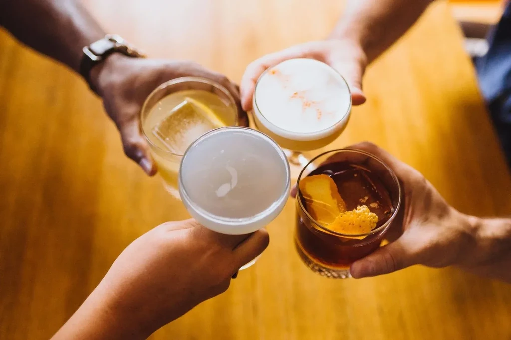 Four diverse pairs of hands toast unique cocktails above wooden table.