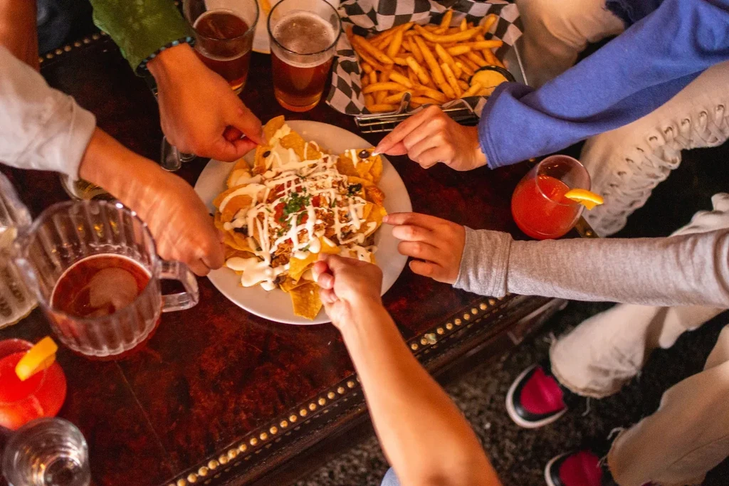 Group sharing loaded nachos, fries, beer, and cocktails on a table.