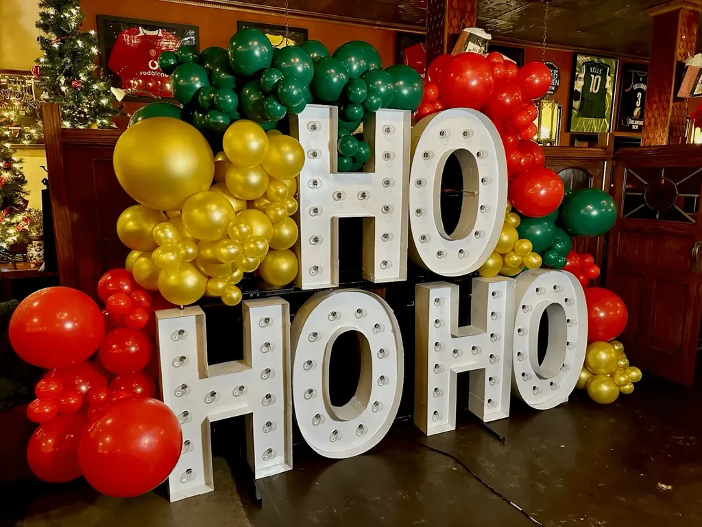 Illuminated 'HO HO HO' marquee letters and festive balloons in a bar.