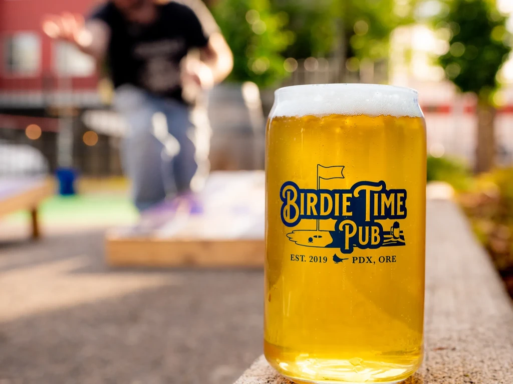 Can-shaped glass of golden beer with "Birdie Time Pub" golf logo outdoors.