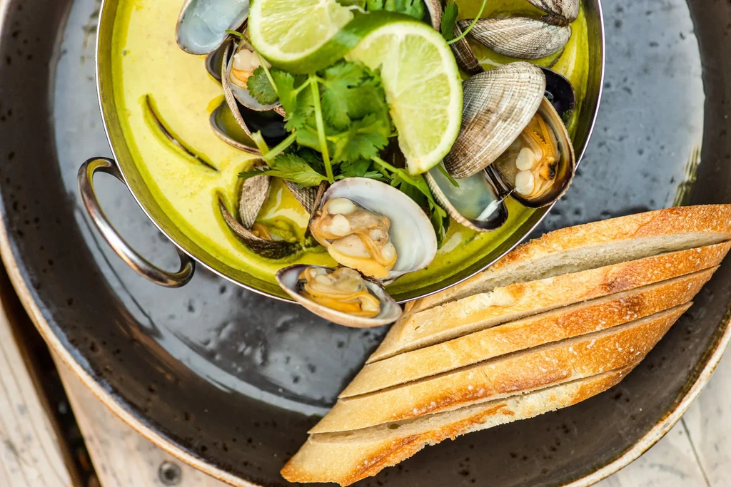 Metal pot of clams in yellow broth with cilantro and baguette.