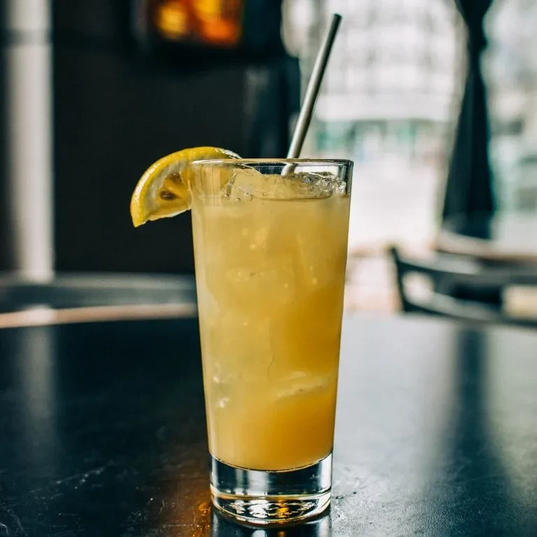 Yellow drink in tall glass with ice, lemon slice, metallic straw.