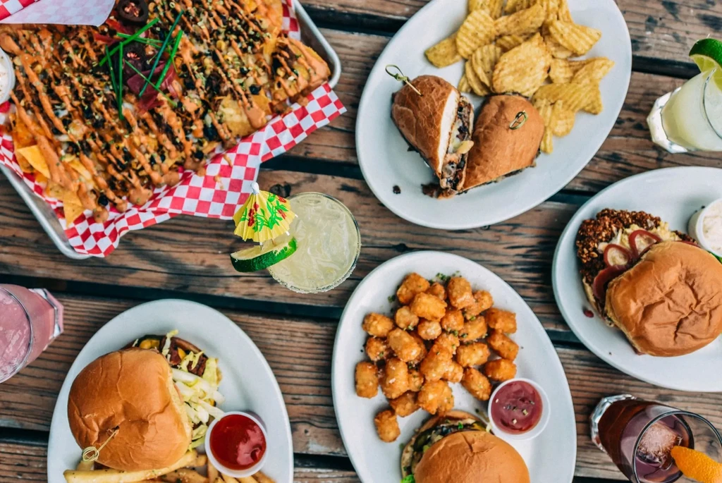 Flat lay: Loaded nachos, burgers, sandwich, tots, cocktails on wooden table.