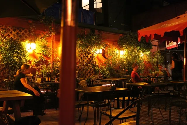 Nighttime outdoor restaurant patio with lush greenery, warm lanterns, and guests.