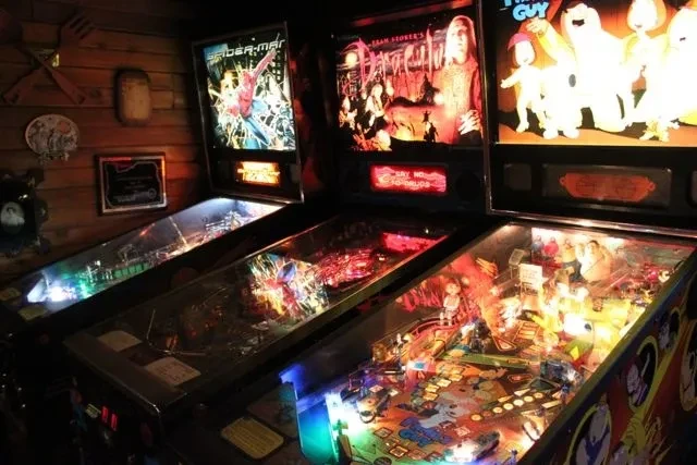 Brightly lit Spider-Man, Dracula, and Family Guy pinball machines in a dark room.