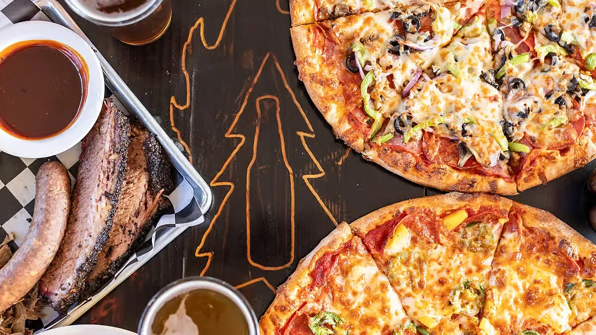 Barbecue tray with brisket and sausage beside two pizzas and beer