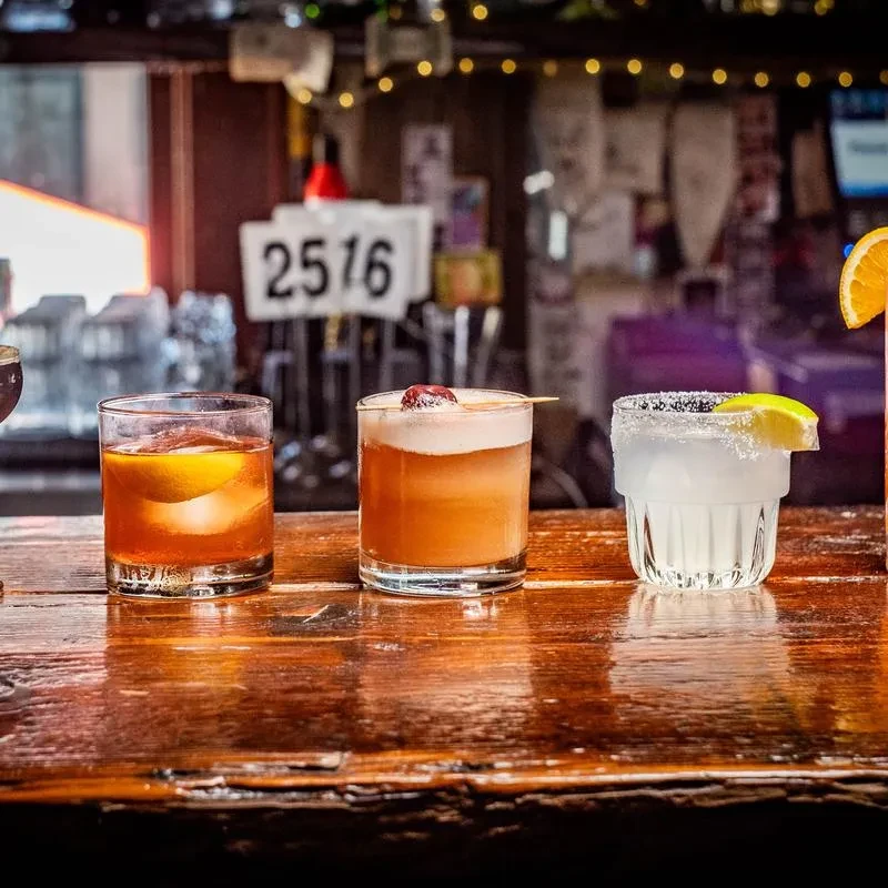 Three distinct cocktails on a dark, reflective bar with blurred lights.