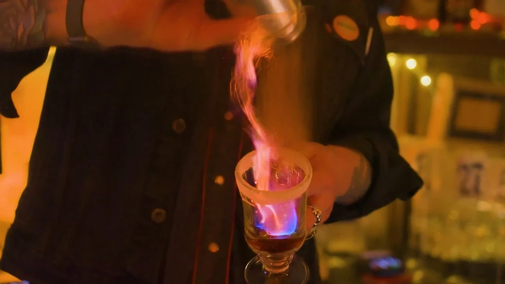 Bartender's hands prepare a flaming cocktail with vibrant orange and blue flames.