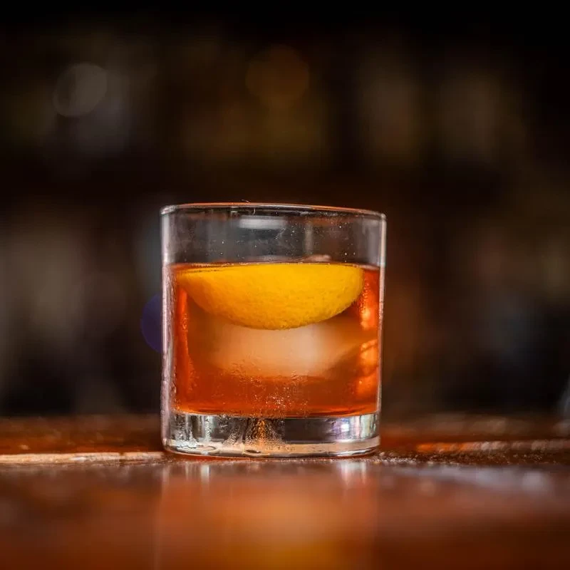 Amber drink with spherical ice and lemon peel on a bar.