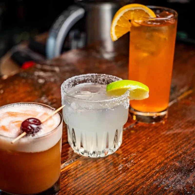Three distinct cocktails with various garnishes on a wooden bar.
