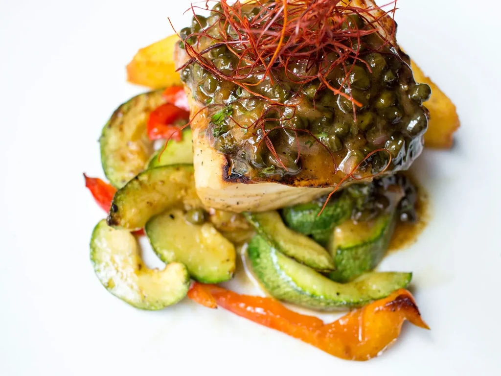 Plated seared fish with green sauce, capers, red spice, zucchini, and potatoes.