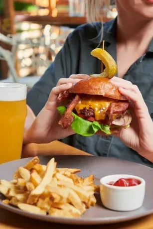 Hands holding an elaborate bacon burger with pickle, fries, ketchup, drink.