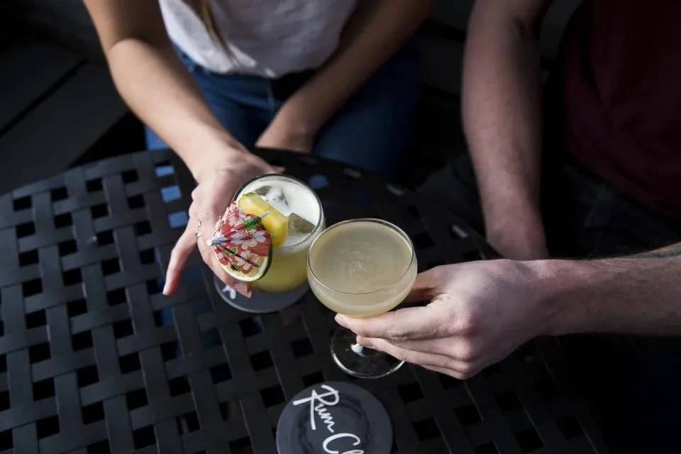 Frothy yellow and clear cocktails held by two hands on a table.