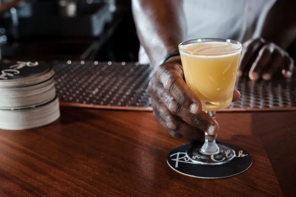 Bartender's hands place a golden cocktail on a "Rum Club" coaster.