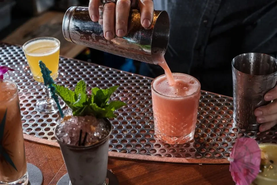 Bartender's hands pour a pink frothy cocktail, surrounded by drinks.