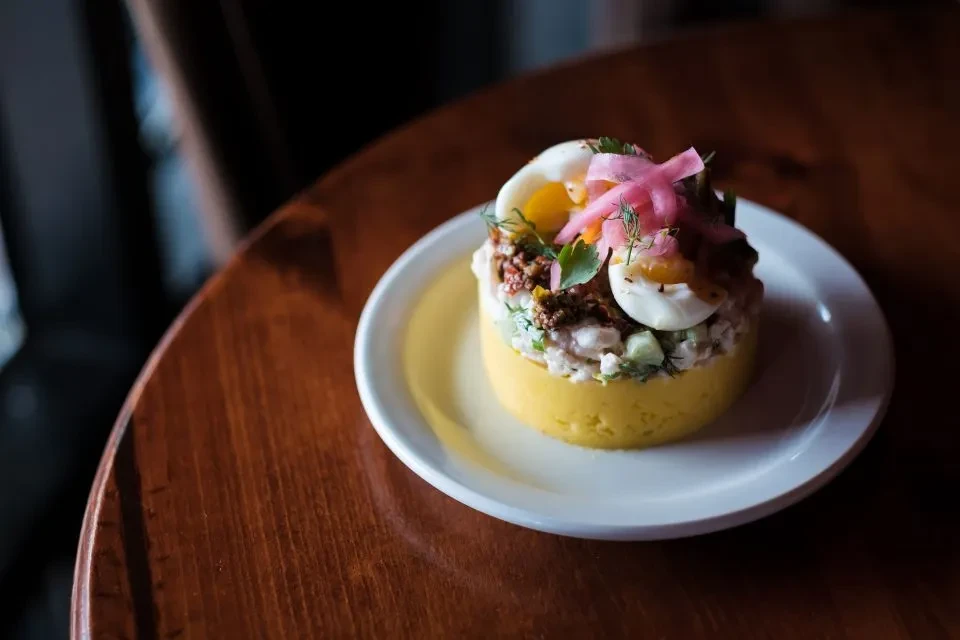 Cylindrical yellow dish plated with eggs, pickled onions, and herbs.