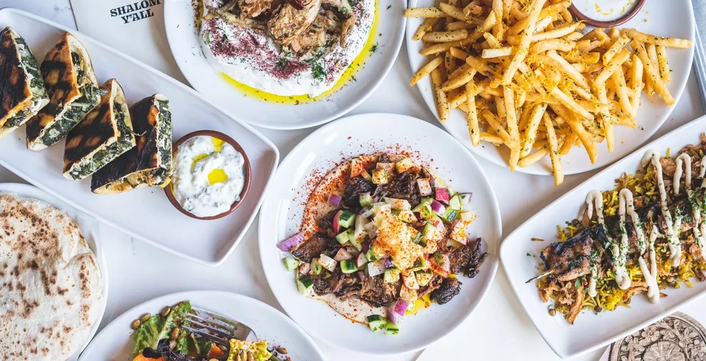 Overhead view of a table with various Mediterranean dishes, including "SHALOM Y'ALL".