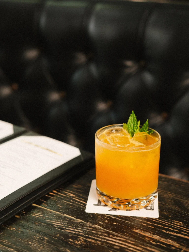 Orange cocktail with mint and ice on wooden table, menu visible.