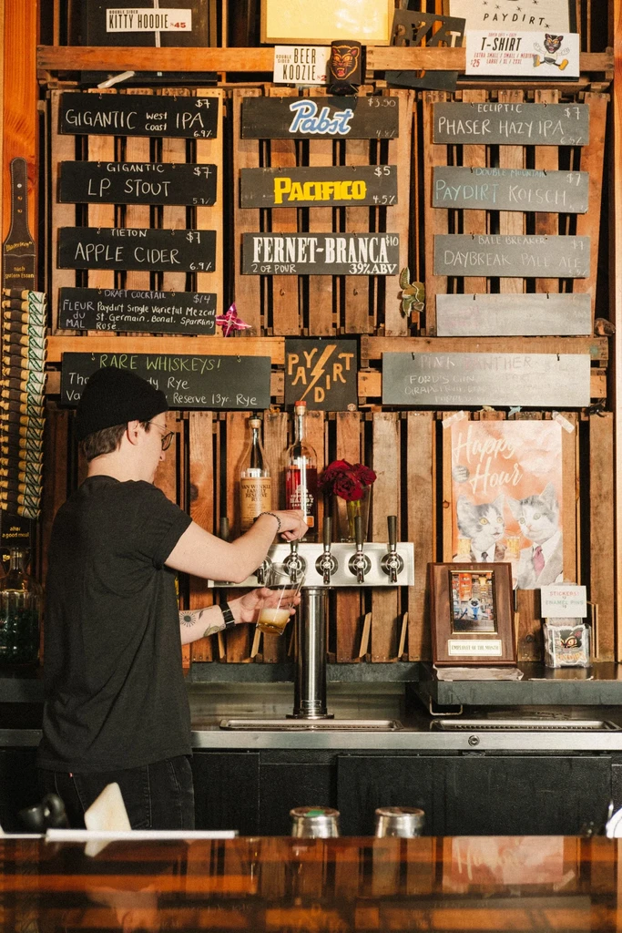 Bartender pours a drink at a bar with a pallet-wall menu display.