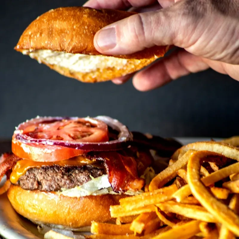Close-up gourmet burger, fries; hand holds top bun for assembly.