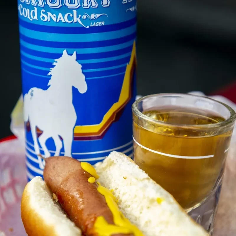 Hot dog with mustard, amber drink, and Cold Snack Lager can.
