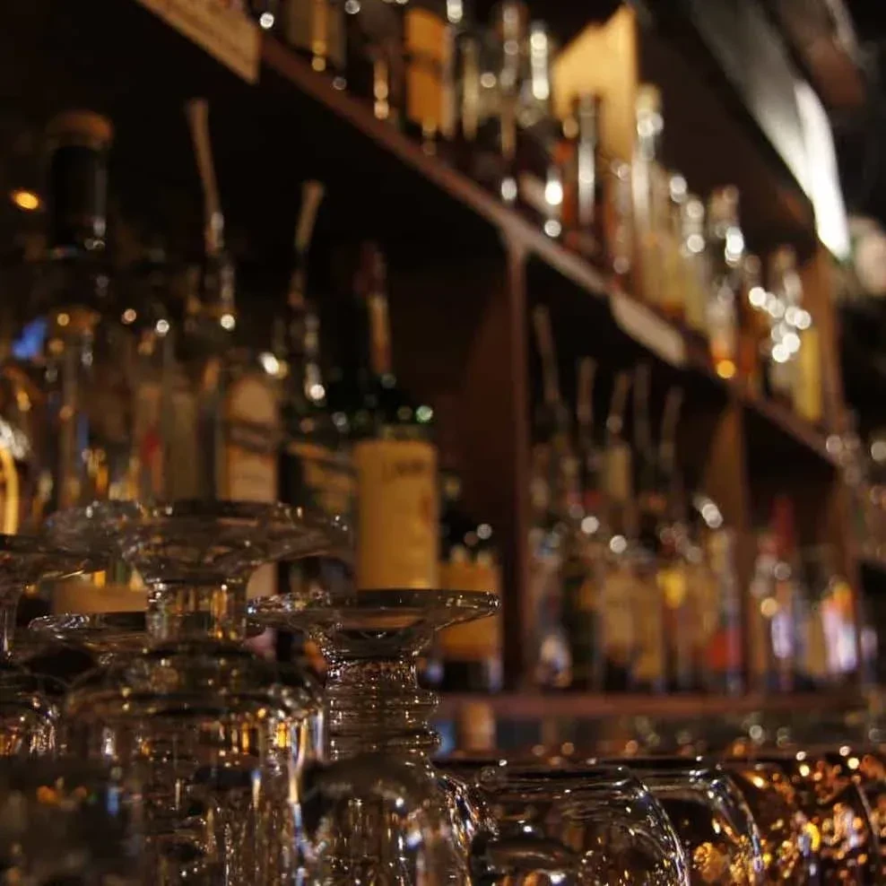 Sharp, inverted drinking glasses in a dim bar, blurred bottles glow.
