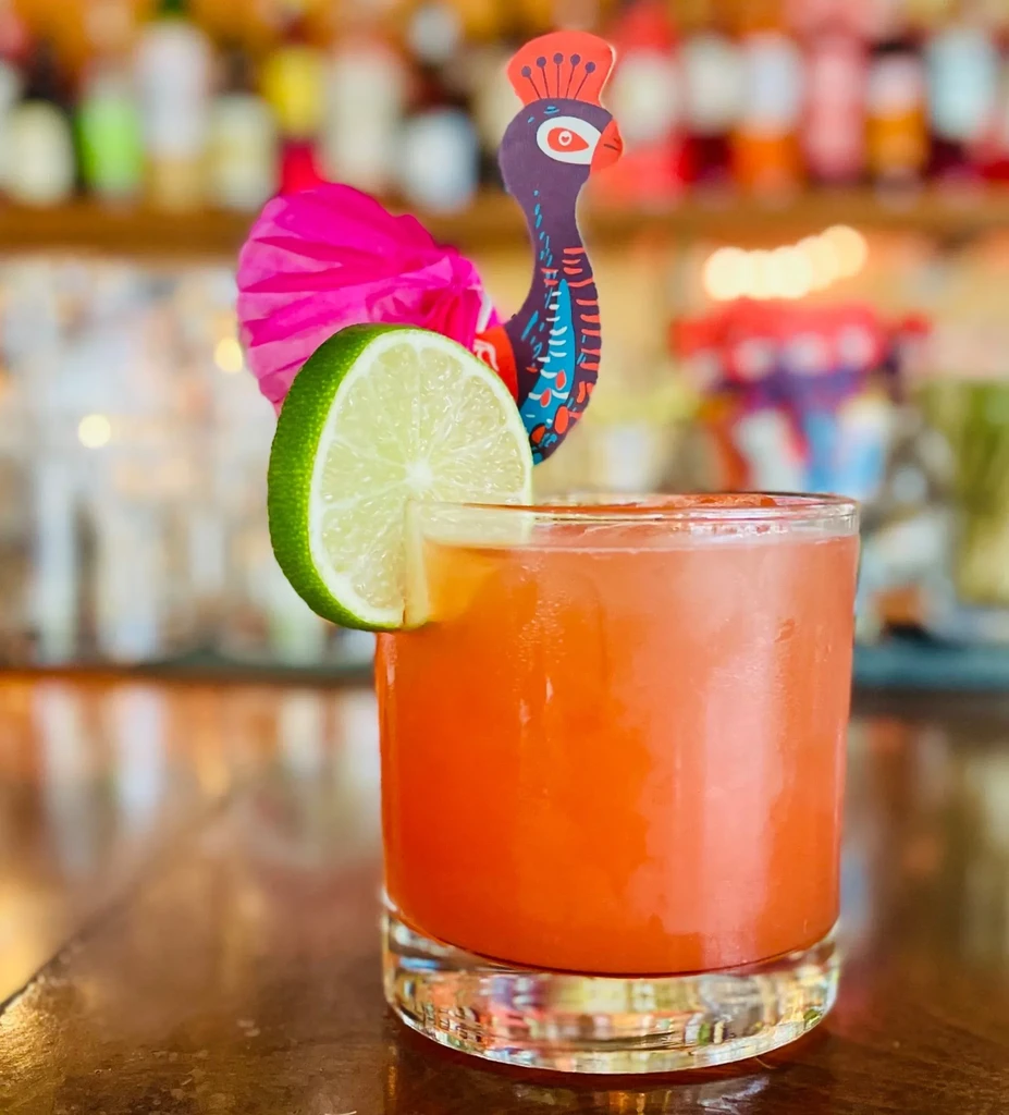 Reddish-orange cocktail with lime and a decorative peacock pick on a bar.