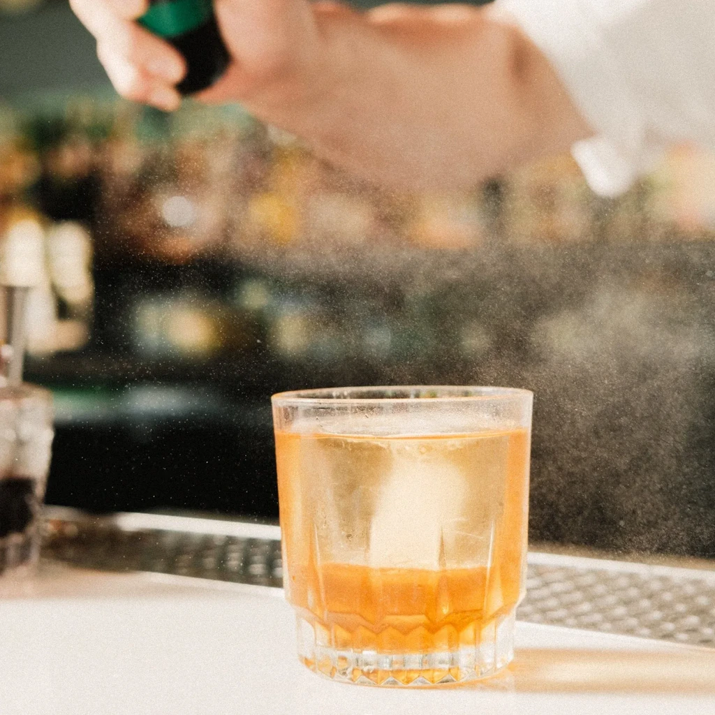 Hand misting an amber drink in a faceted glass on a bar.