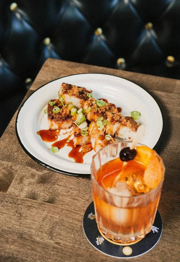 Fried pork belly dish and amber cocktail on a rustic wooden table.