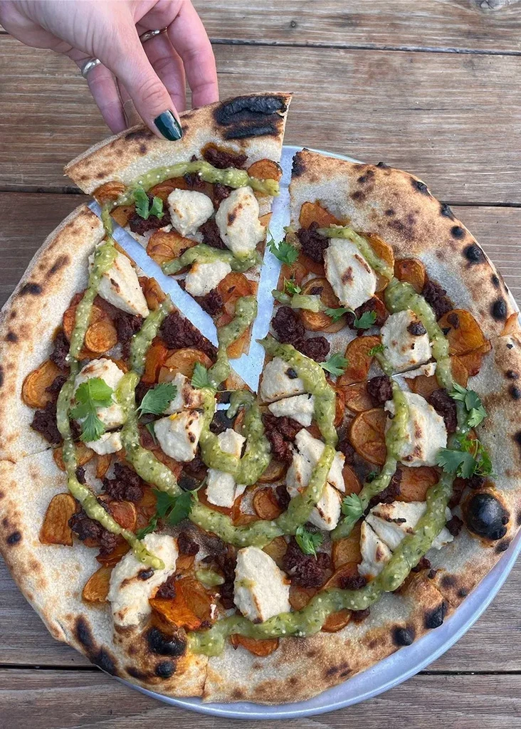 Hand lifts colorful pizza slice with diverse toppings and green sauce.
