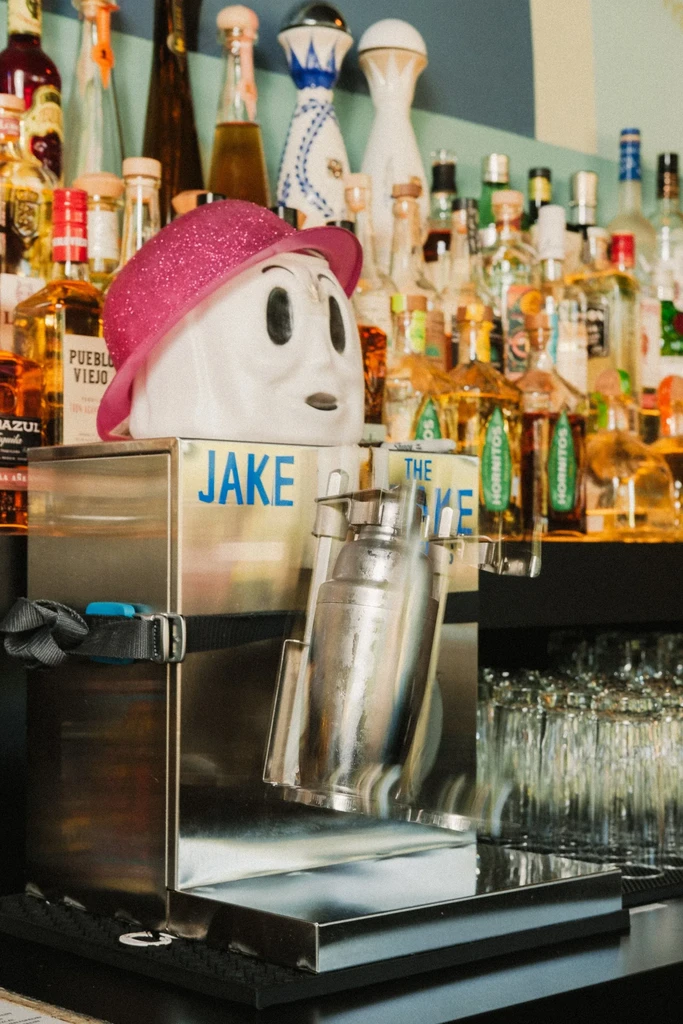 Frozen drink machine, cartoon head, pink fedora, shaker, and bar bottles.