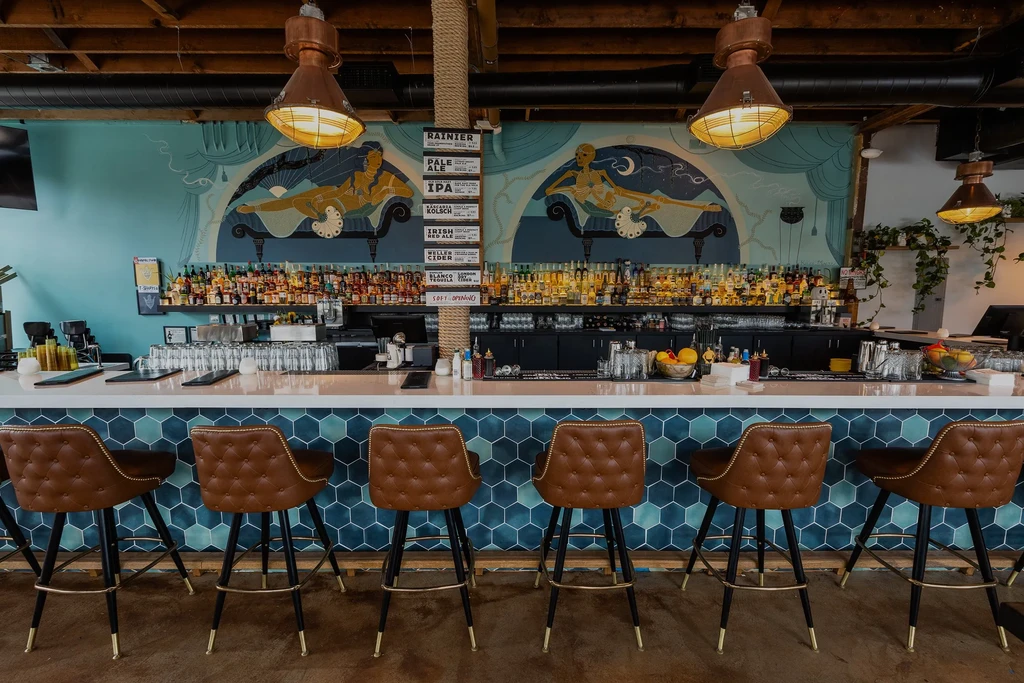 Empty bar with tufted stools, teal tiles, liquor, and a mural.