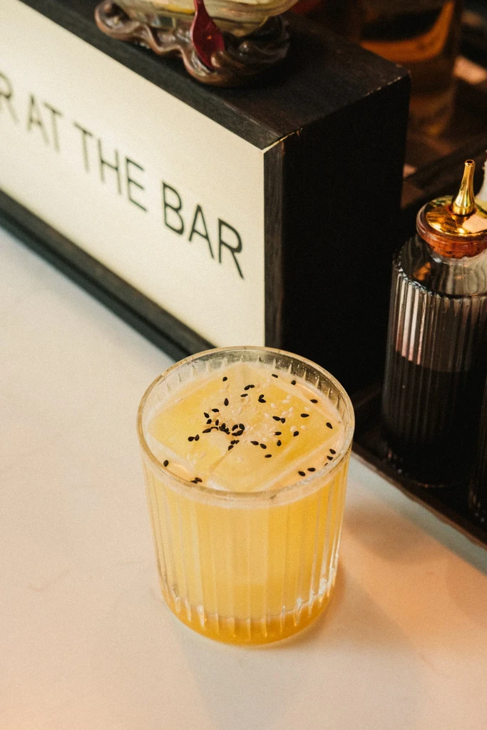 Close-up: Light yellow cocktail with ice, sesame seeds, and white crystals.
