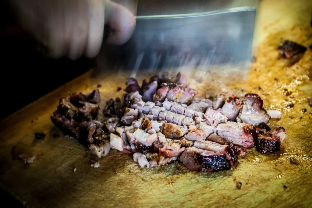 Hand chops cooked meat with a blurred cleaver on wooden board.