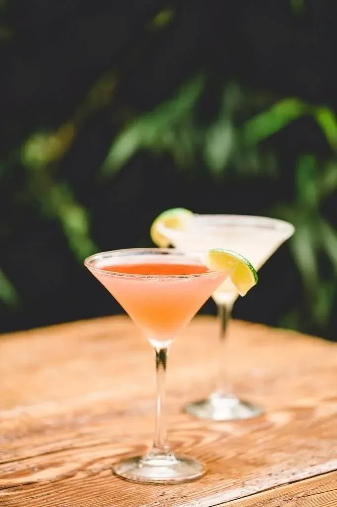 Two lime-garnished cocktails on a wooden surface, with blurred green background.