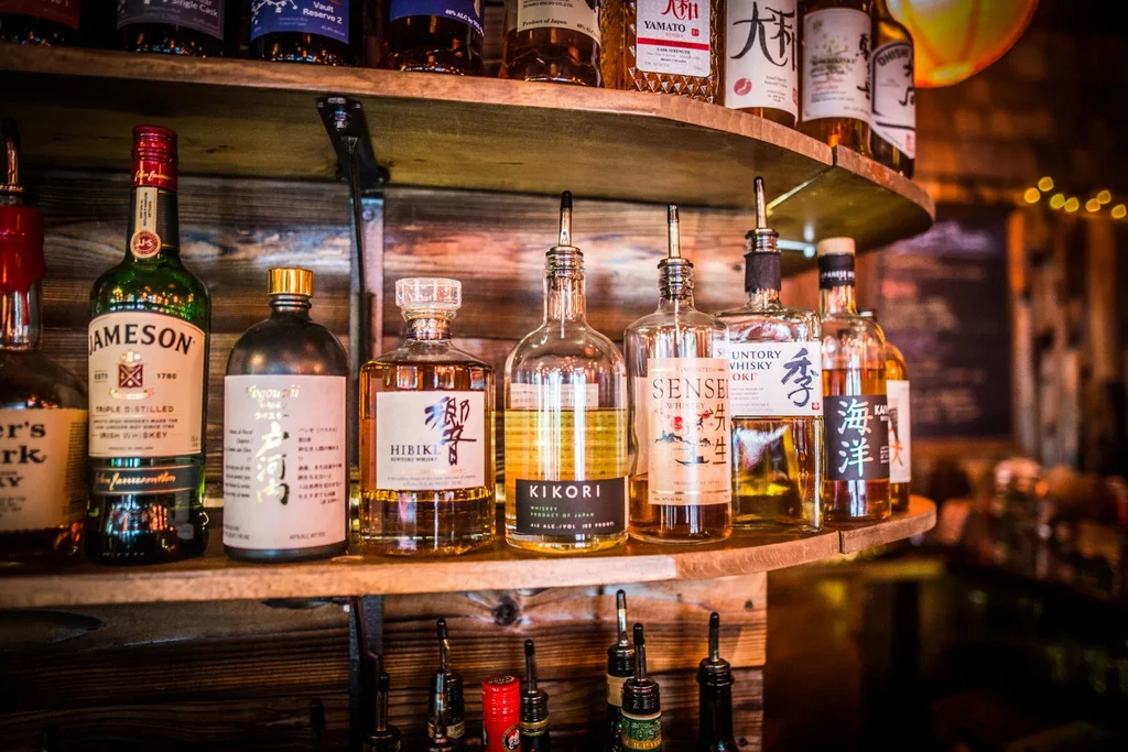 Whiskey bottles with pour spouts on rustic wooden bar shelves.